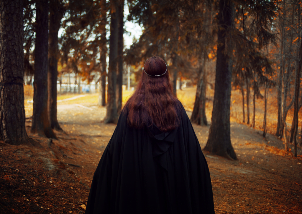 The back of a woman walking through a wood wearing a black cloak and looks a bit like a witch. 