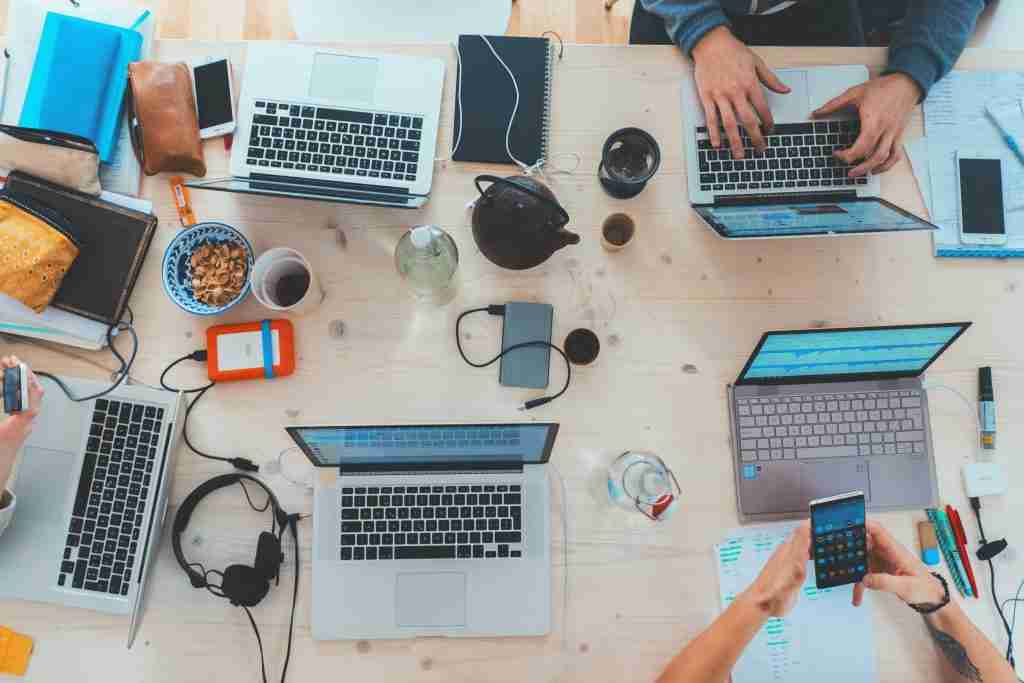 Image of a table with 5 laptops and mobiles on it as well as drinks and people taking notes